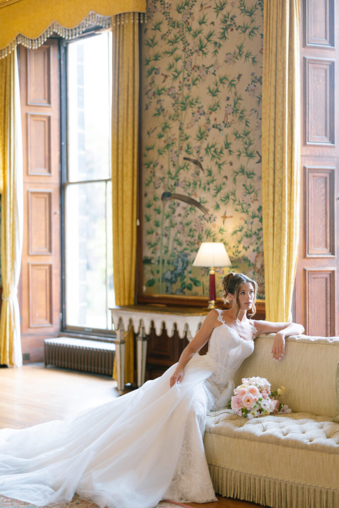 A bride in a white gown sits elegantly on a cream-colored sofa, holding a bouquet of flowers, in a luxurious room with tall wooden windows, gold curtains, and floral wallpaper.
