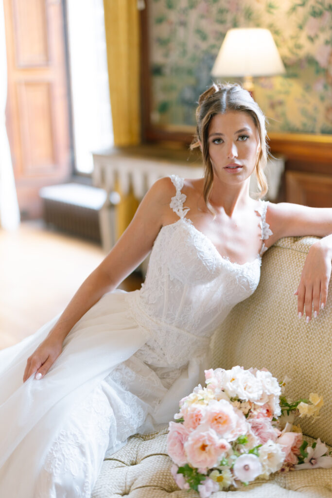 A woman in a white lace wedding dress sits on a cream-colored sofa, next to a bouquet of pink and white flowers, in a softly lit, elegant room with floral wallpaper and a lamp in the background.