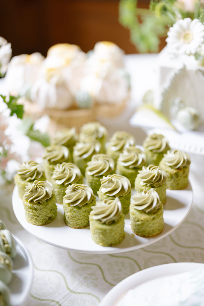 A plate of small, round green cakes topped with swirls of frosting, arranged neatly on a white stand. The background features other desserts and floral decorations on a white tablecloth.
