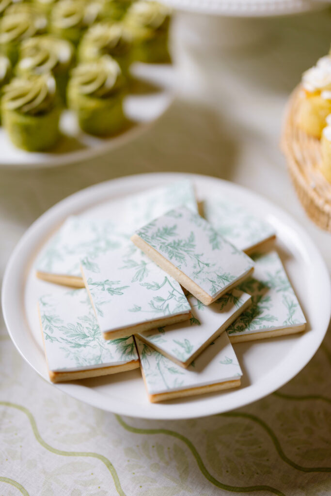 A white plate holds rectangular cookies decorated with white icing and green botanical patterns. In the background, there are green frosted cupcakes and part of a basket of yellow treats.