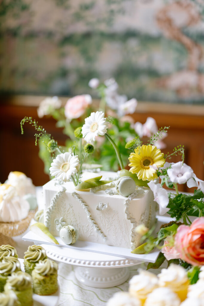 A white, rectangular cake decorated with green leaves, yellow and white flowers, and green macarons sits on a cake stand, surrounded by floral arrangements and other green and yellow pastries.