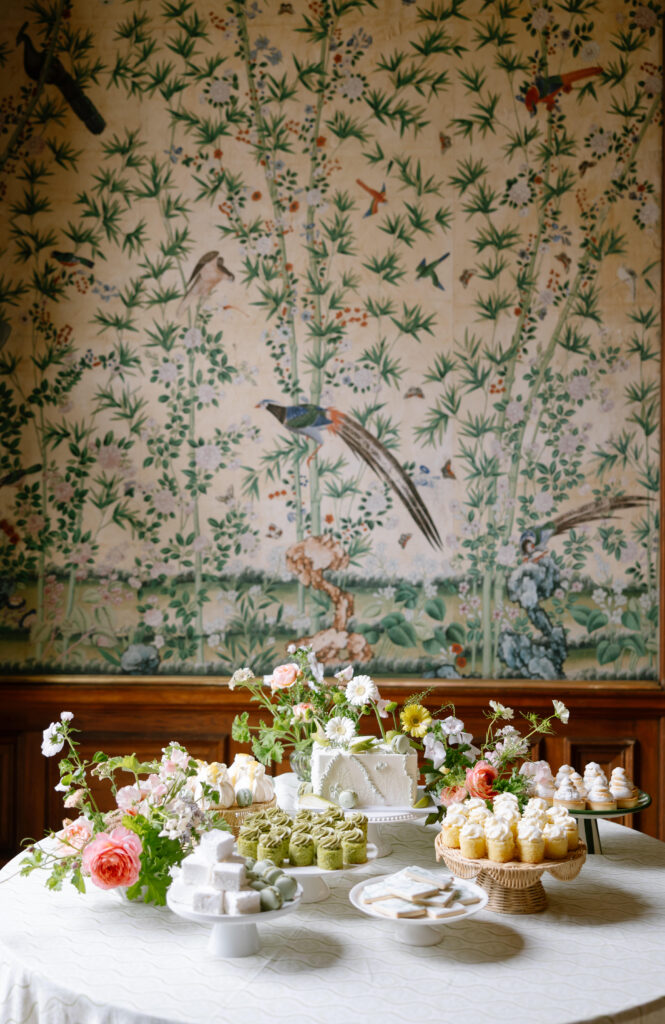 A round table with assorted pastries, cakes, and floral arrangements sits in front of a wall decorated with a detailed bird and bamboo mural.