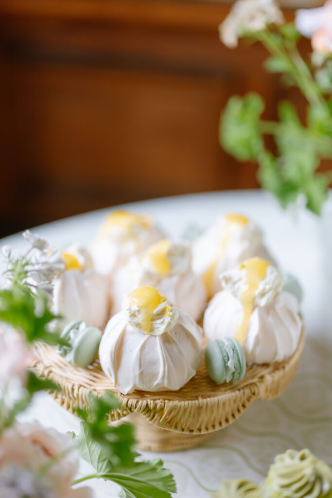 A wicker plate holds white meringue desserts topped with yellow cream and surrounded by green macarons on a table, with blurred flowers and greenery in the foreground.