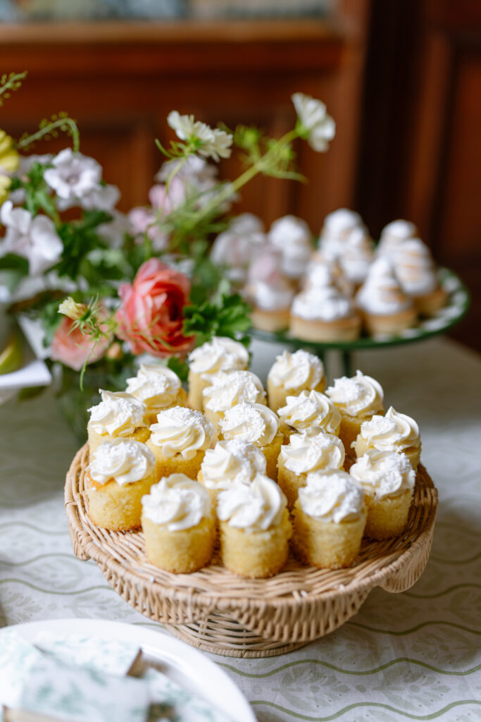 A wicker tray filled with small yellow cakes topped with swirls of white cream sits on a table, with more decorated cupcakes and flower arrangements in the background.