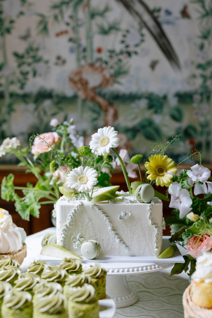 A white rectangular cake decorated with green leaves, white and yellow flowers, and green macarons sits on a white cake stand, surrounded by other floral desserts on a table with a botanical background.