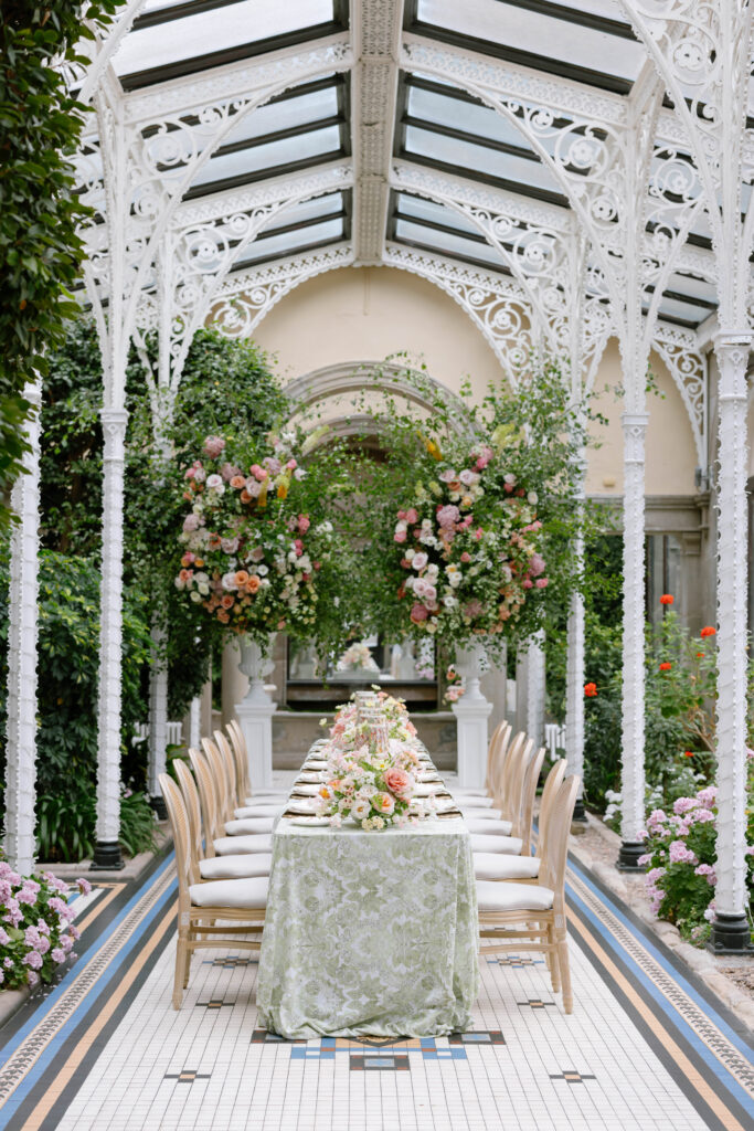 A long, elegant dining table with floral arrangements set under a glass-roofed, ornate white conservatory, surrounded by lush greenery and blooming flowers.