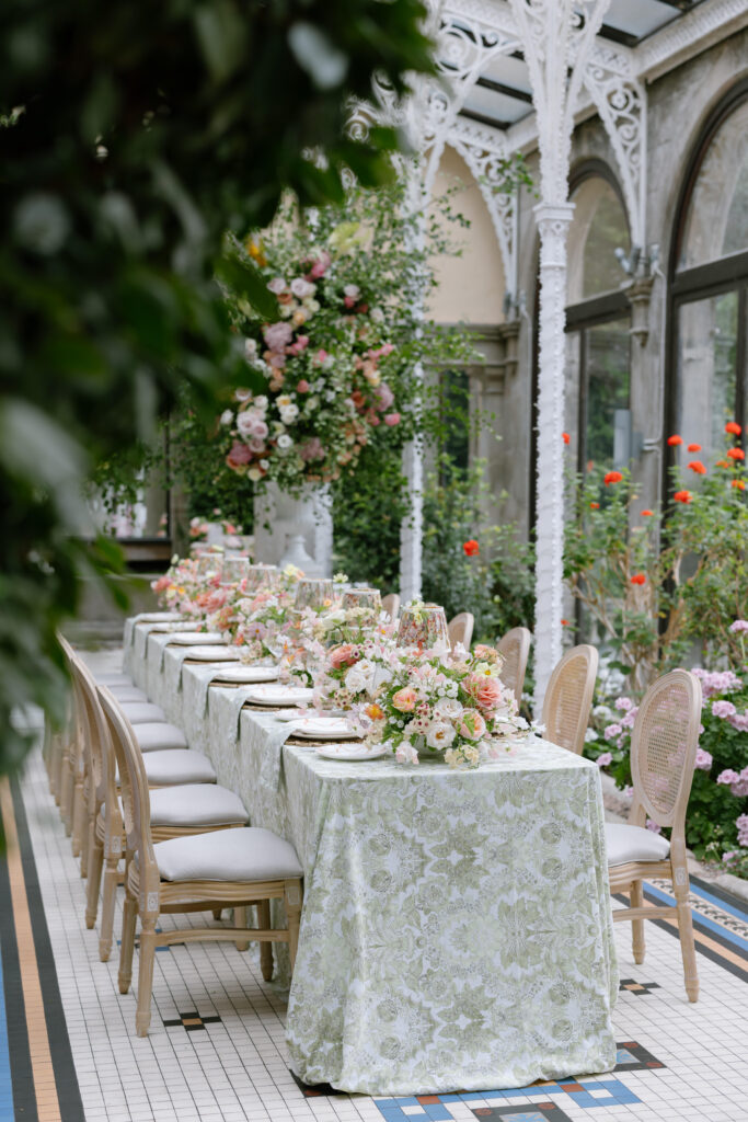 A long, elegant table with floral-patterned tablecloth, set for a meal with pastel flower arrangements and beige chairs, sits in a glass conservatory surrounded by lush greenery and colorful blooms.