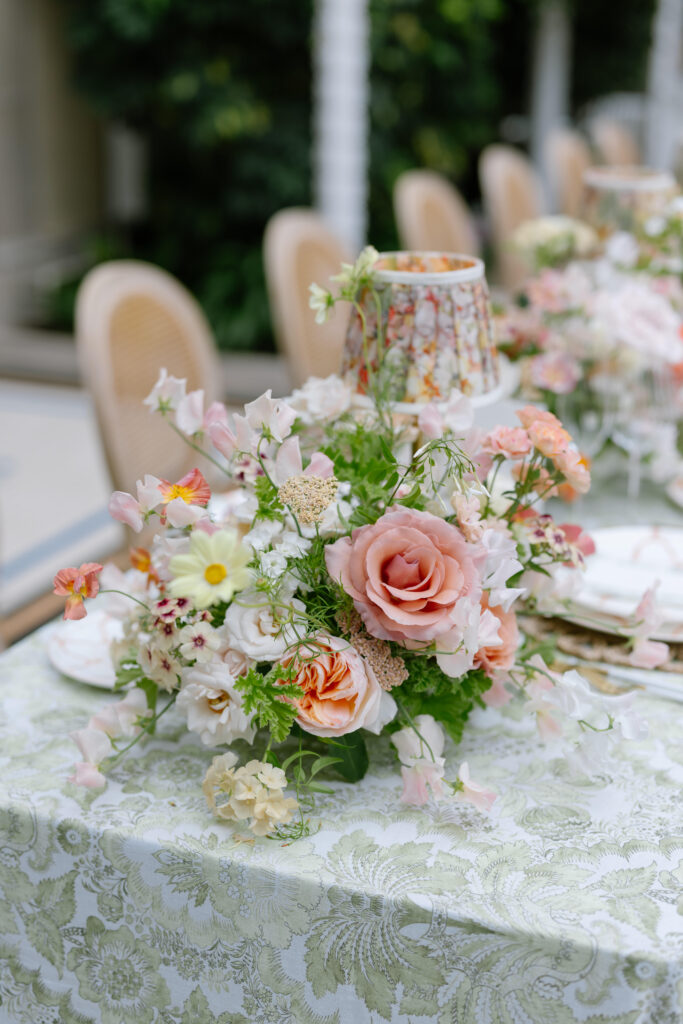 A close-up of an elegant outdoor table setting with a floral tablecloth, a bouquet of pink and peach roses, white flowers, greenery, and a small decorative lamp in the center. Wicker chairs are in the background.