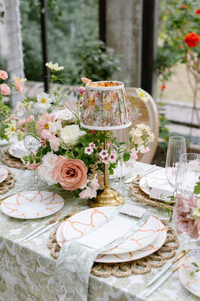 A beautifully set table with floral arrangements, elegant plates, gold cutlery, woven placemats, glassware, and a small decorative lamp with a floral shade, surrounded by greenery and blooming flowers.
