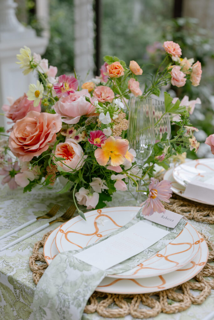 Elegant table setting with a white plate featuring orange accents, a floral napkin, gold cutlery, and a place card. A vibrant bouquet of pink, peach, and yellow flowers is arranged in a glass vase nearby.