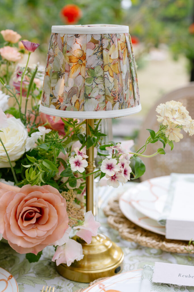 A close-up of an elegant table setting with a brass lamp featuring a floral-patterned shade, surrounded by pink and white flowers, and a plate with a name card on a woven placemat in the background.