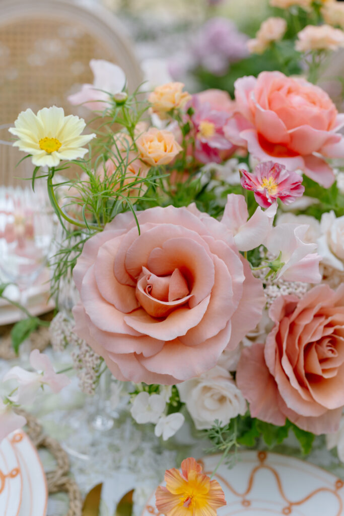 A close-up of a floral arrangement featuring large blush pink roses, peach-colored flowers, white blossoms, and greenery, set on a table with elegant dishware in the background.