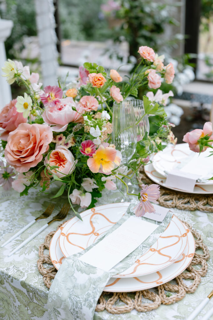 A close-up of an elegant table setting with floral-patterned plates, a woven placemat, a printed menu, green and white napkins, glassware, and a centerpiece of pink and peach flowers with greenery.