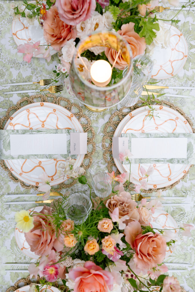 A beautifully set table with floral-patterned plates, gold accents, elegant menus, and lush arrangements of pink and peach flowers surrounding a lit candle centerpiece.