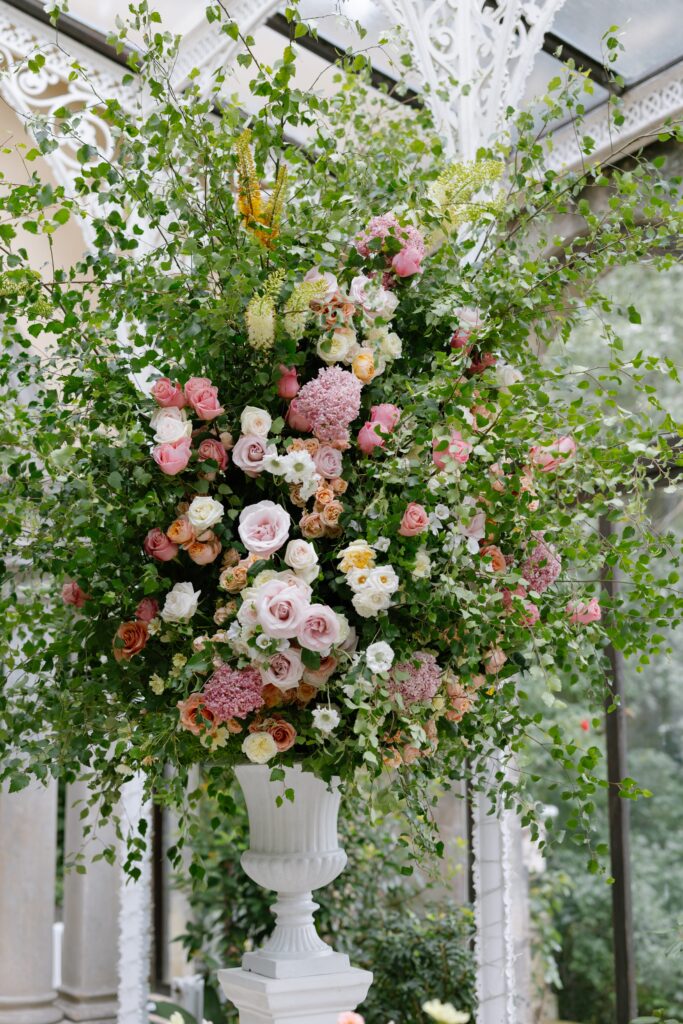 A large, ornate white vase overflows with a lush floral arrangement of pink, white, and yellow roses, hydrangeas, and abundant greenery, set in a bright, elegant conservatory.