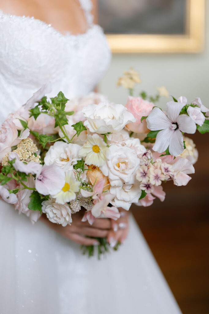 A bride in a white lace wedding dress holds a bouquet of pastel flowers, including white, pale pink, and cream blooms, with green foliage. The background is softly blurred.