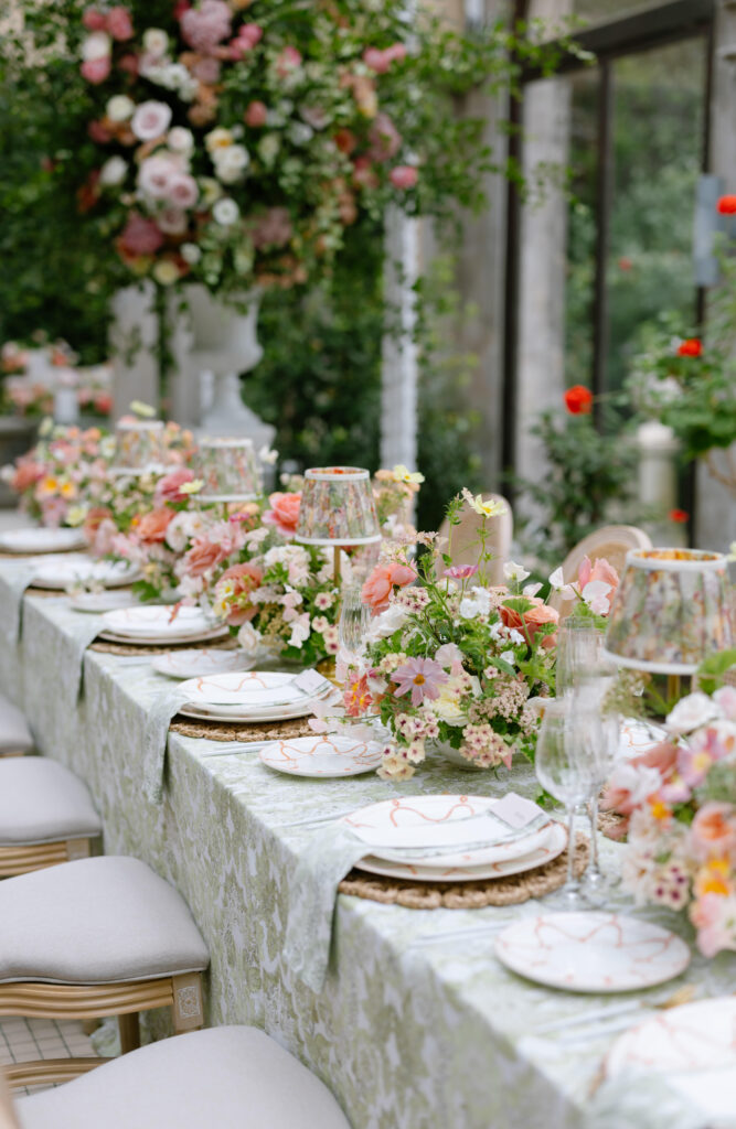 An elegant outdoor table is set with white plates, floral-patterned napkins, and glasses. The table is decorated with lush bouquets of pink and peach flowers, and matching lamps, creating a romantic, garden-party atmosphere.