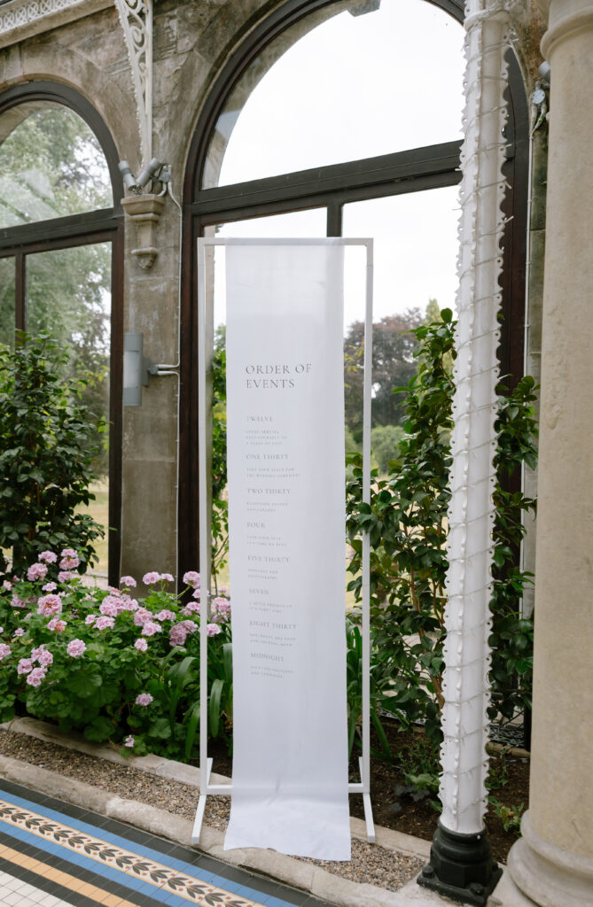 A tall white sign with “Order of Events” and a schedule printed in elegant text stands indoors by large windows, with pink hydrangeas and greenery visible outside. The setting appears formal and bright.
