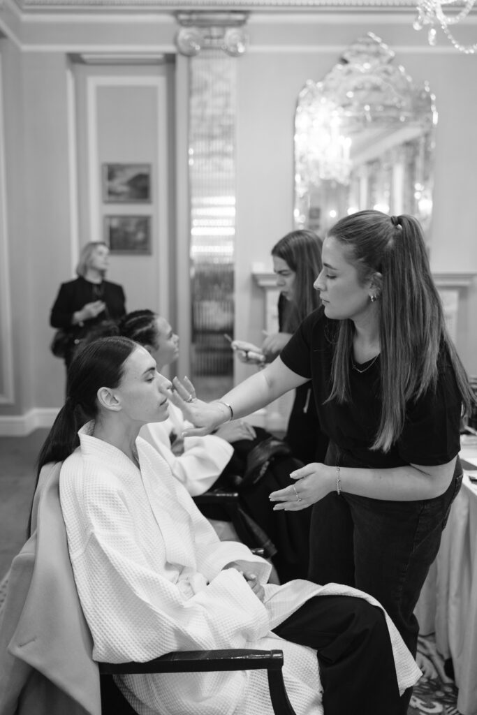 A makeup artist applies makeup to a seated woman in a robe. Two other women sit nearby, also receiving makeup. The setting is an elegant room with ornate decor, mirrors, and soft lighting.