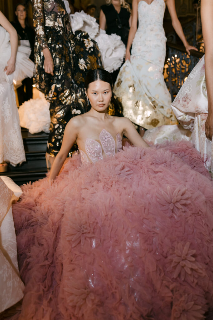 A model in a voluminous pink gown adorned with large floral appliqués sits on a staircase, surrounded by other models in elegant, patterned dresses. The setting appears to be an ornate, indoor event.