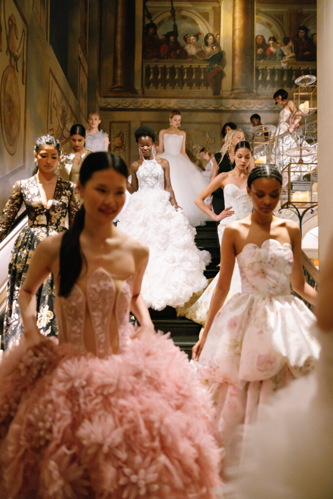 A group of models in elaborate, elegant gowns descend a grand staircase in an ornate, classical interior with painted walls and soft lighting, creating a luxurious and sophisticated atmosphere.
