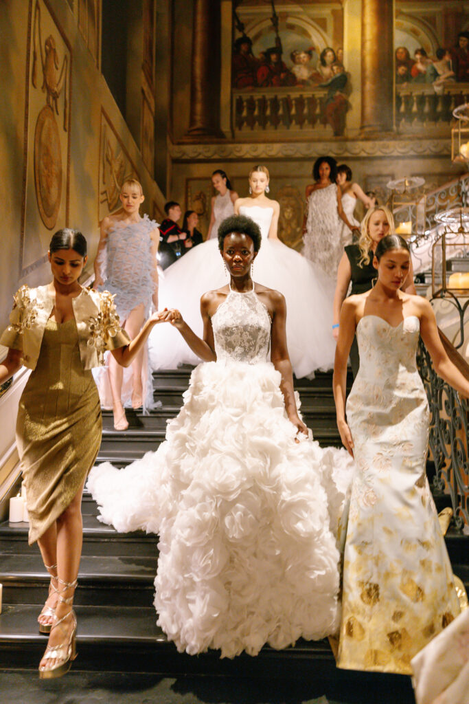 A group of models in elegant, high-fashion gowns descend a grand staircase in a lavish, ornately decorated setting, with the central model wearing a voluminous white dress adorned with floral textures.