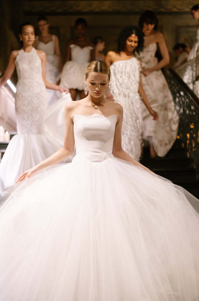 A bride in a strapless white ballgown descends a staircase, followed by several women in elegant white dresses, all in a softly lit, formal setting.