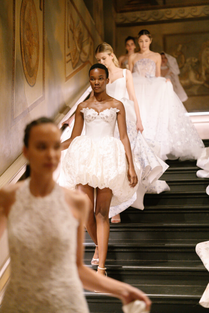 Models wearing elegant white dresses descend a grand staircase with ornate wall paintings, showcasing intricate bridal or formal gowns in a fashion show setting.