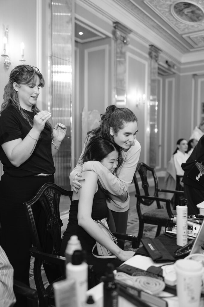 Two women share a warm hug while sitting at a makeup station in an elegant room. Another woman stands nearby, smiling and holding a makeup brush. The atmosphere is lively and joyful.