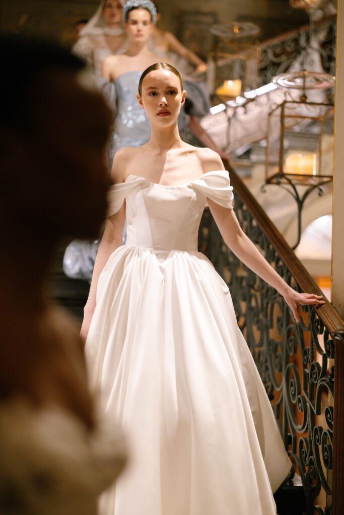A woman in an elegant off-the-shoulder white gown stands on a grand staircase, holding the railing, with other people in formal attire visible in the background.