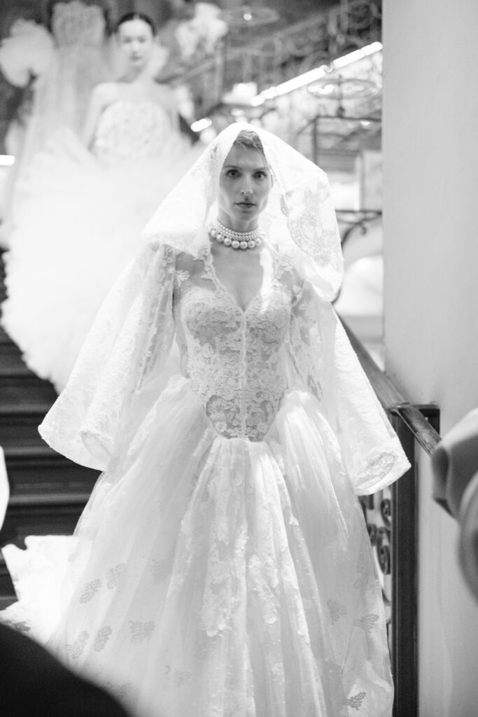 A person in an ornate lace wedding dress and veil, wearing a pearl necklace, walks down a staircase. Other people in similar dresses are visible in the background. The image is in black and white.