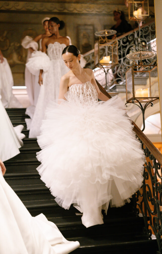 A model in a voluminous, white, ruffled gown descends a staircase, followed by others in elegant white dresses, in a grand setting with ornate railings and warm candlelight.