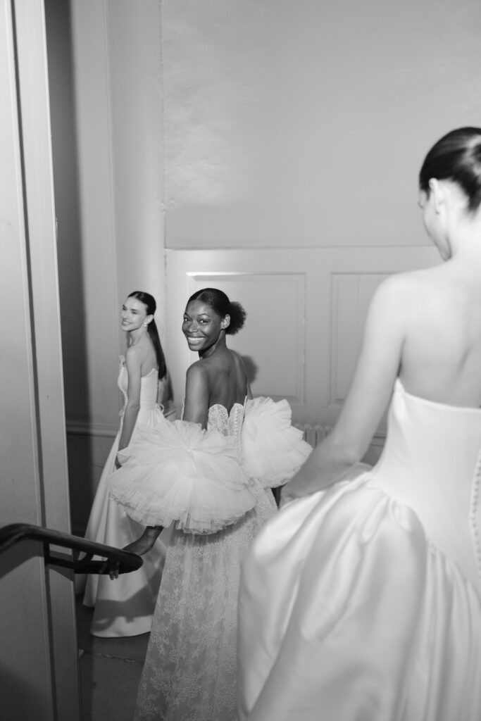 Three women in elegant, strapless white gowns smile and walk down a hallway. The image is black and white, and the mood is joyful and celebratory, as if they are backstage at a fashion show or wedding.