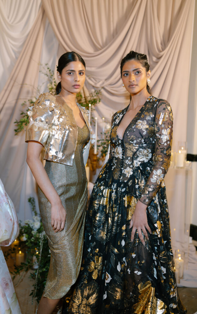 Two women pose in elegant evening gowns. One wears a metallic gold dress with a short jacket, the other wears a long black and gold floral dress. Both stand against a draped fabric backdrop with candles and greenery.