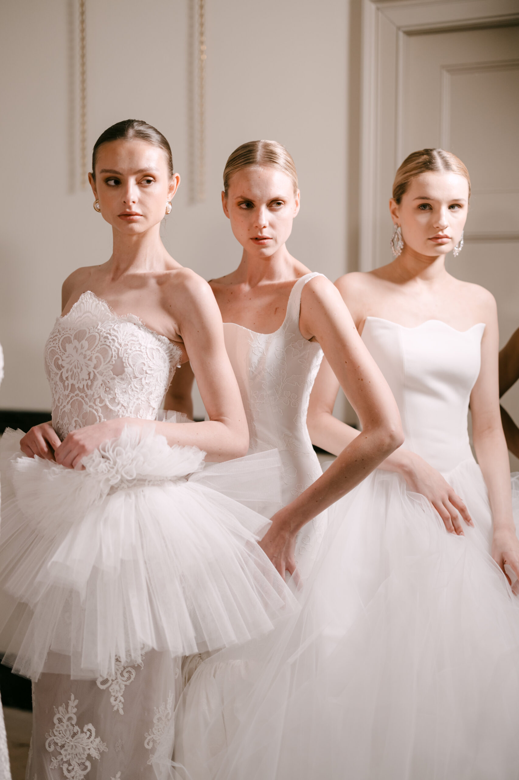 Three women stand side by side wearing elegant white wedding dresses with different styles and textures, including lace and tulle, in a well-lit indoor setting.
