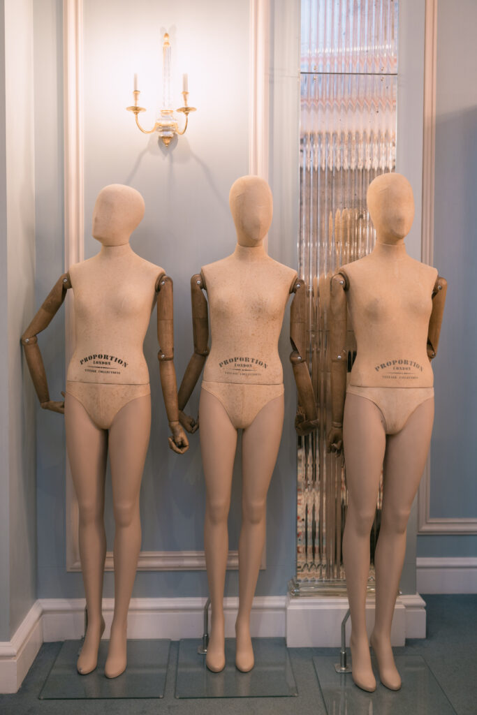 Three faceless mannequins with wooden arms stand side by side in a well-lit room with elegant wall paneling and a sconce above them. The word “PROPORTION” is printed on their torso areas.