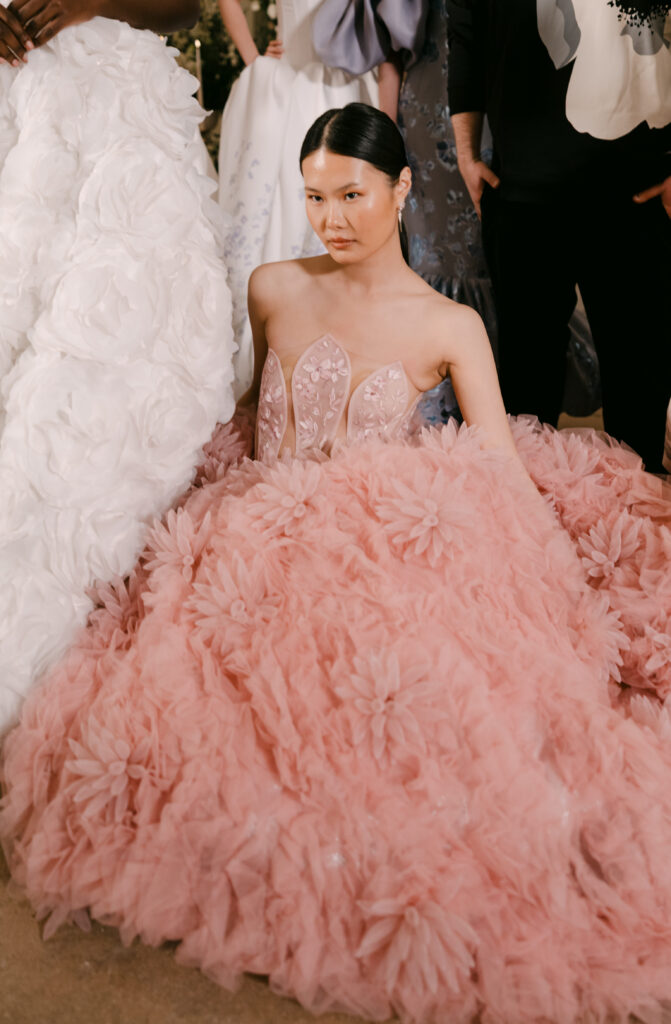 A woman with sleek, dark hair sits on the floor wearing a strapless, pale pink gown adorned with intricate floral details and a voluminous, textured skirt. People in elegant attire stand around her.