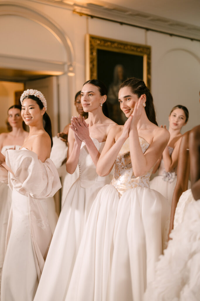 A group of women in elegant white gowns stand together indoors, smiling and clapping. The setting appears formal, with soft lighting and a framed portrait in the background, suggesting a celebratory or ceremonial event.