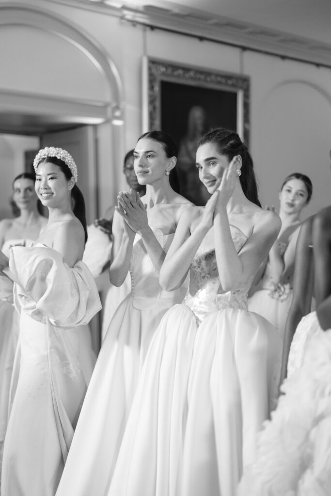 A group of models in elegant, flowing white gowns stand together indoors, smiling and clapping. The image is in black and white, with a classic, celebratory atmosphere.