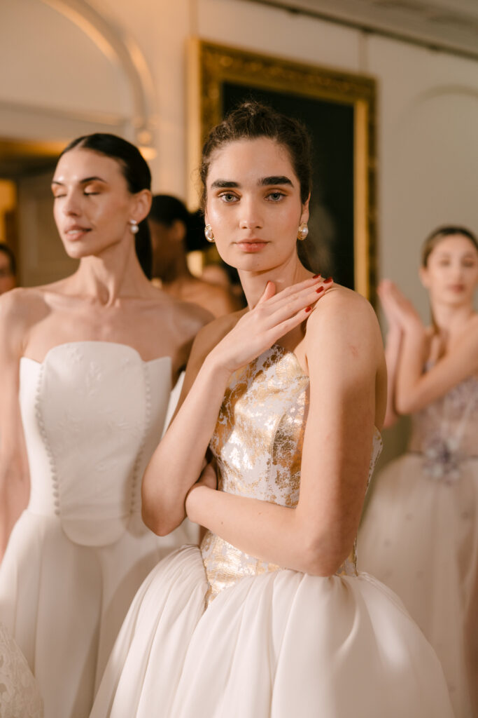 A young woman in an elegant gold and white gown poses with her hand on her shoulder among other women in white dresses, all standing indoors in a softly lit, formal setting.
