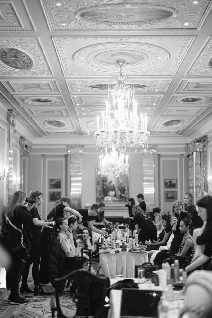 A black-and-white photo of a grand, ornate room with a chandelier and decorated ceiling, where multiple people are getting their hair styled by stylists at several tables. The scene appears busy and professional.