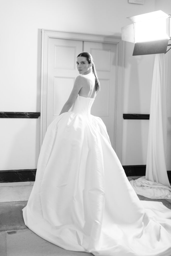 A woman in a strapless, voluminous white gown stands in front of a closed door, looking over her shoulder. The scene is lit by a large studio light, creating a dramatic, elegant atmosphere.