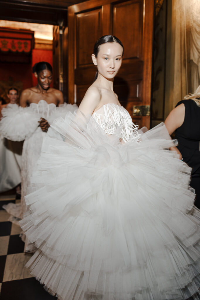 Two models in elaborate white tulle gowns stand indoors, with one looking at the camera. The dresses are voluminous and dramatic, featuring layered ruffles. The setting appears elegant with checkered flooring and wooden doors.
