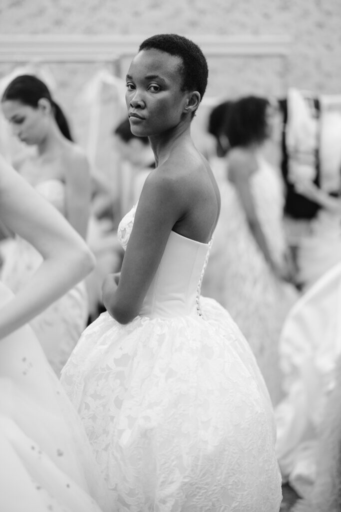 A woman in a strapless, textured white gown stands among others in similar dresses. She looks over her shoulder toward the camera in a softly lit, elegant setting. The image is in black and white.