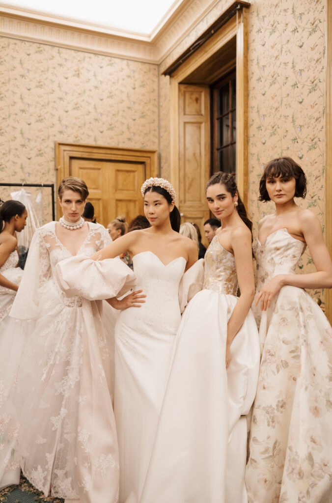 Four models wearing elegant, white and cream floral wedding gowns pose together indoors, standing close in front of a wooden door and patterned wallpaper.