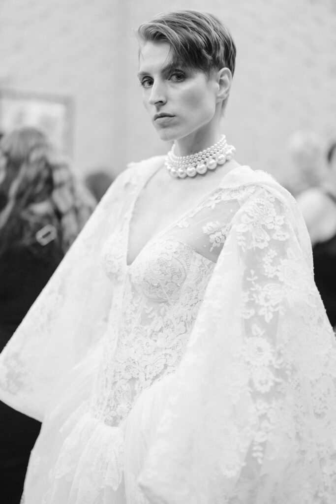A person with short hair wears an elegant lace wedding dress with dramatic sleeves and a layered pearl necklace, looking confidently at the camera in a softly focused, indoor setting.