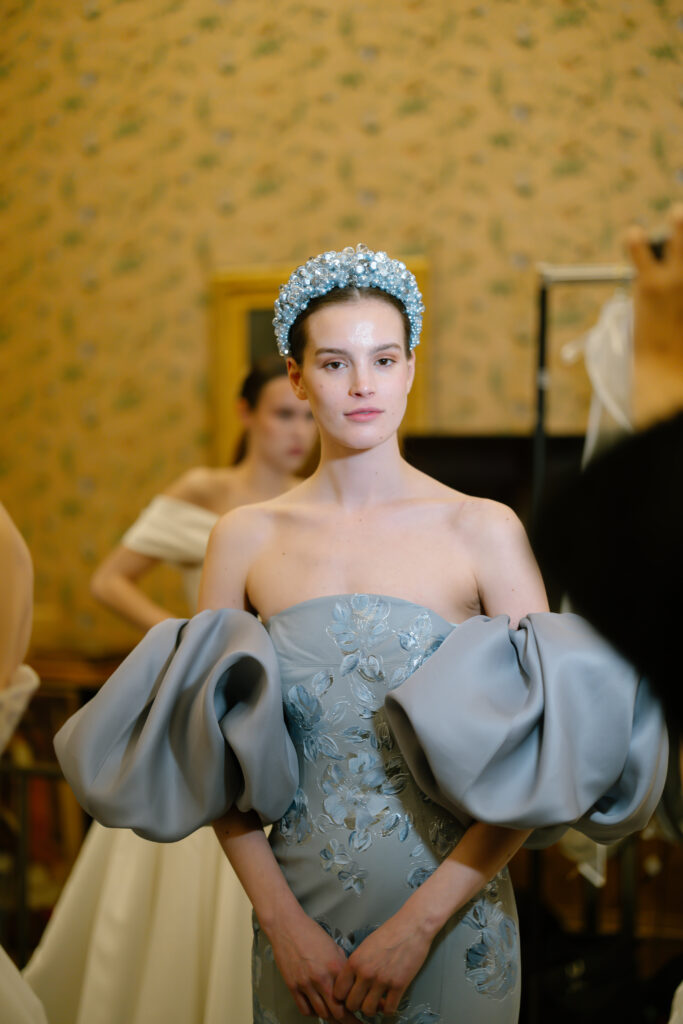 A woman wearing a light blue, off-the-shoulder gown with large puff sleeves and floral embroidery stands indoors. She has a blue beaded headband and looks at the camera, with another person blurred in the background.