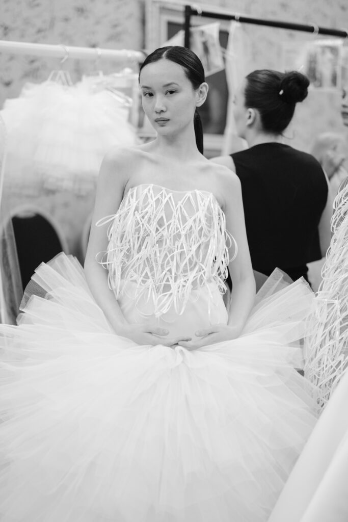 A model in a white, avant-garde dress with a textured bodice and voluminous tulle skirt stands backstage, looking at the camera, with another person visible in the softly focused background.