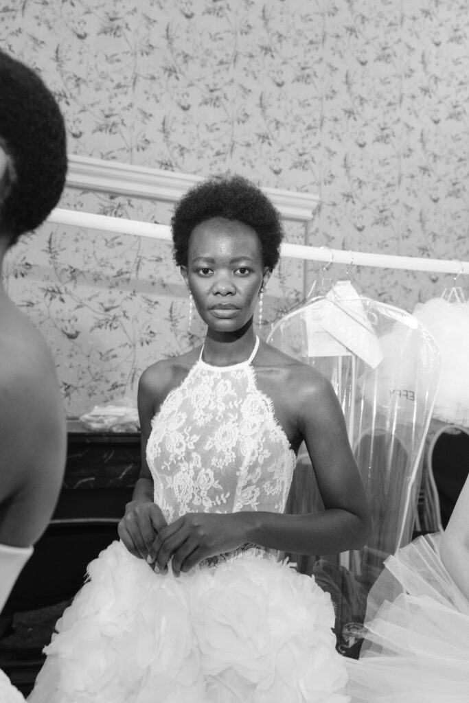 A woman wearing a lace bodice and tulle skirt stands in a dressing room, looking at the camera. Garment bags and other outfits hang in the background, and she is surrounded by others preparing for an event. The photo is in black and white.