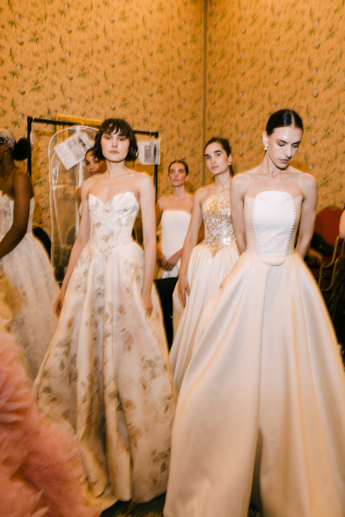 Four women wearing elegant, floor-length strapless gowns stand together in a softly lit room with floral wallpaper, preparing for a fashion show or event. The atmosphere is calm and anticipatory.